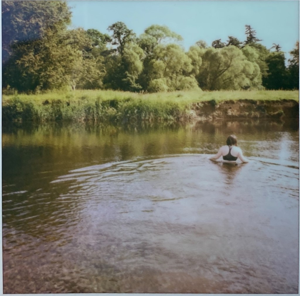 colour polaroid of a woman half-submerged in the river, the sky is clear blue and the banks are green.